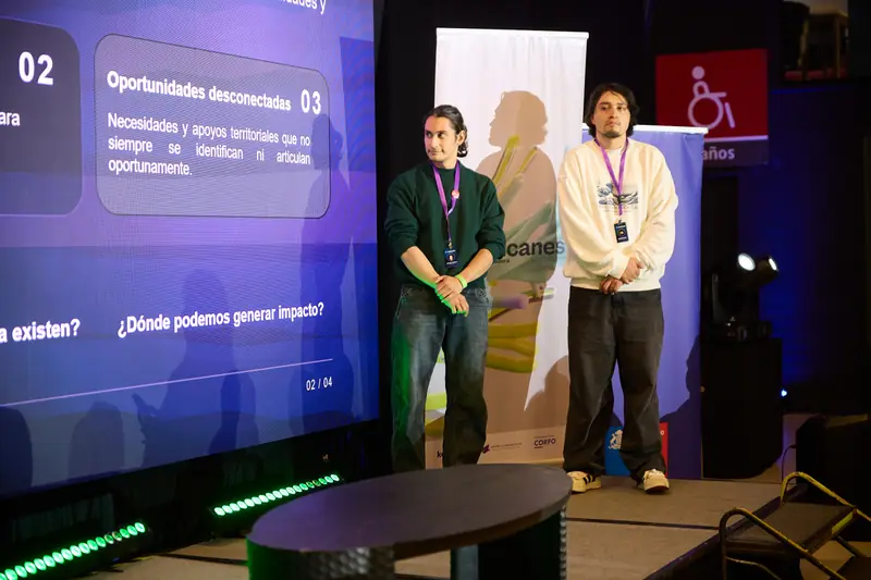 Dos integrantes del equipo en el escenario junto a la lámina “Oportunidades desconectadas” y la pregunta “¿Dónde podemos generar impacto?”.