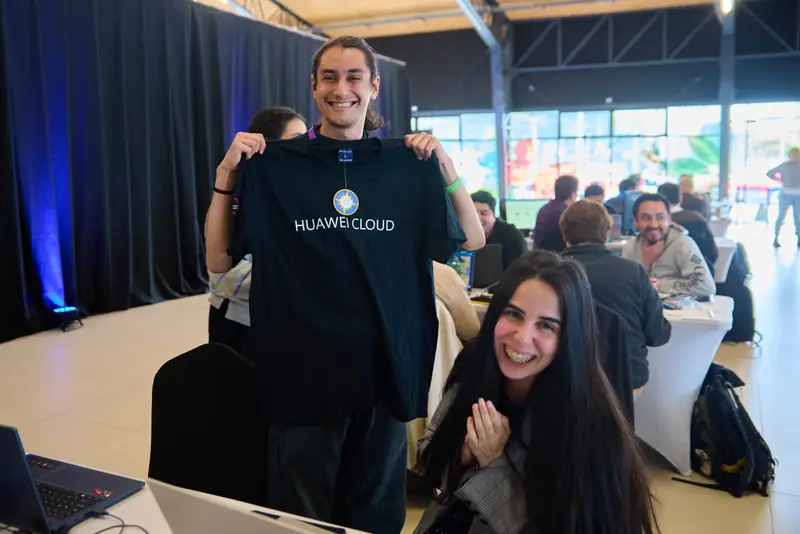 Un integrante del equipo sonríe sosteniendo una polera de Huawei Cloud; a su lado, otra integrante celebra sentada a la mesa.